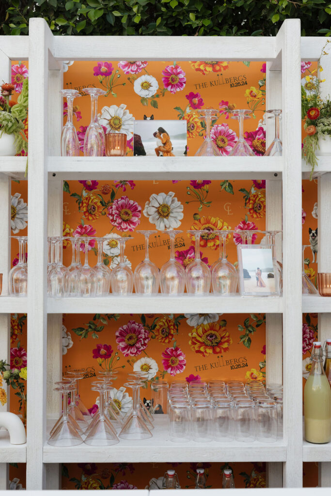 Backlit shelves with glassware and floral backdrop - Shelves filled with glassware in front of a bright floral backdrop featuring "The Kullbergs" logo and wedding date, with small decorative elements.