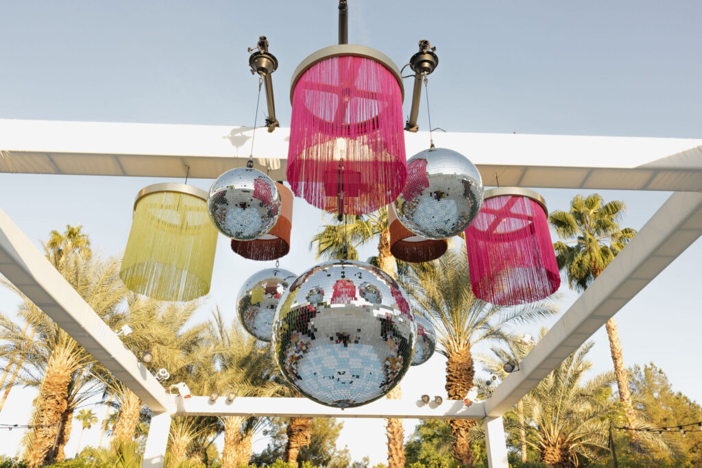 A festive display of disco balls and colorful fringe lanterns hanging above a long reception table setup, creating a lively atmosphere in an outdoor setting.