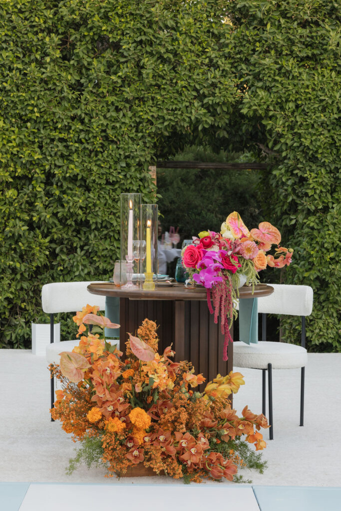 A sweetheart table adorned with large floral arrangements in shades of orange and pink, two candle holders, and surrounded by a lush green hedge backdrop.