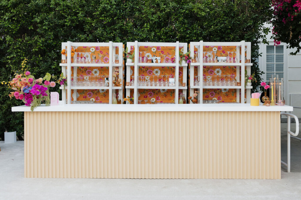 Photo of a beautifully decorated outdoor bar setup - A stylish bar setup with a bright floral backdrop, glassware, and pink and orange flower arrangements displayed against a lush green hedge at the Park Palm Springs Hotel.