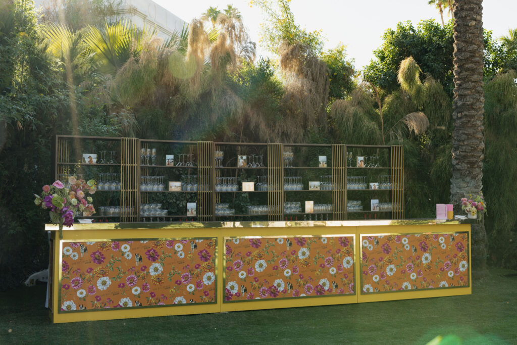 A beautifully designed gold bar with floral panels at Park Palm Springs Hotel, decorated with glassware and small framed photos, blending into the lush garden setting.