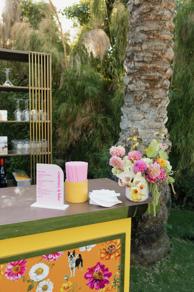 Colorful floral-themed bar setup with a pink drink menu and straw holder - A floral-patterned bar with a colorful arrangement of pink and yellow flowers, drink menus, and pink straws, set against a palm tree at Park Palm Springs Hotel.