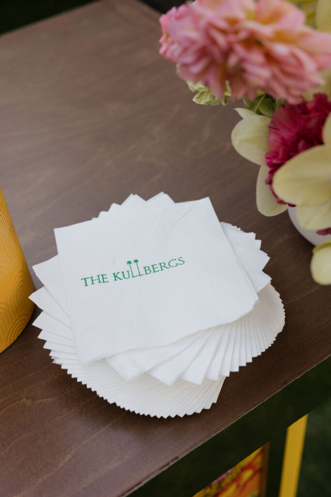 A close-up of white napkins printed with “The Kullbergs” logo, placed on a wooden table beside a floral arrangement.