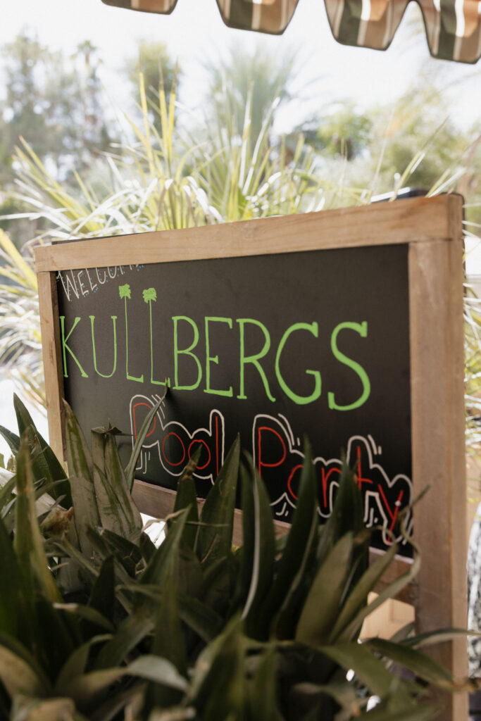 A chalkboard sign reading "Kullbergs Pool Party" stands among tropical plants, inviting guests to a poolside celebration at Park Palm Springs Hotel.