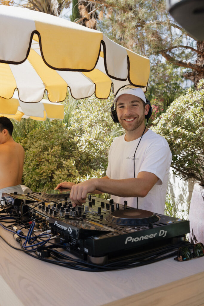 A DJ smiling while mixing music outdoors at Park Palm Springs Hotel, under a yellow-and-white striped umbrella, providing a fun vibe for the celebration.