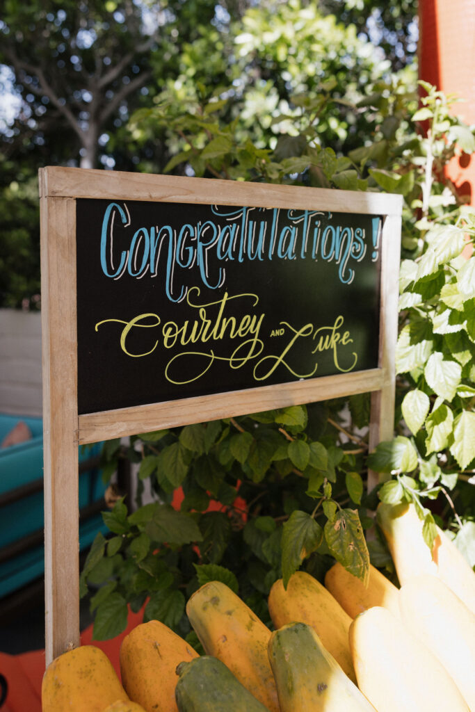 A chalkboard sign with "Congratulations Courtney and Luke" written in elegant script, surrounded by greenery and fruit, welcoming guests to the event.