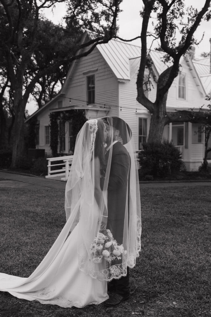 A romantic black-and-white moment of the bride and groom kissing under the bride's lace veil, with The Winfield Inn's historic house in the background.