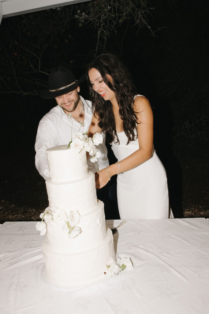 The bride and groom laugh as they cut their three-tiered white wedding cake adorned with fresh white flowers under the evening sky.