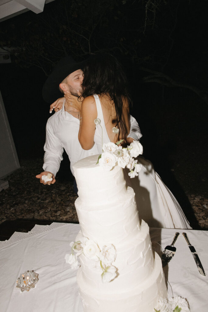 The bride and groom kiss passionately during the cake-cutting moment, with a tiered white cake adorned with fresh floral accents in the foreground.