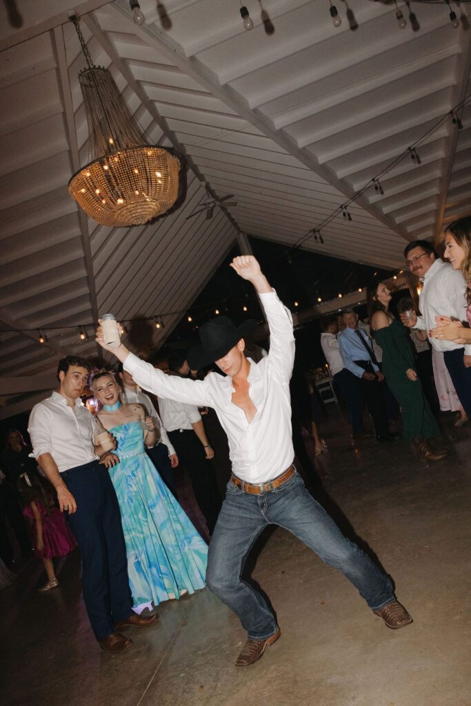 A cowboy-hat-wearing guest enthusiastically dancing in a white shirt and jeans under twinkling string lights at the wedding reception.