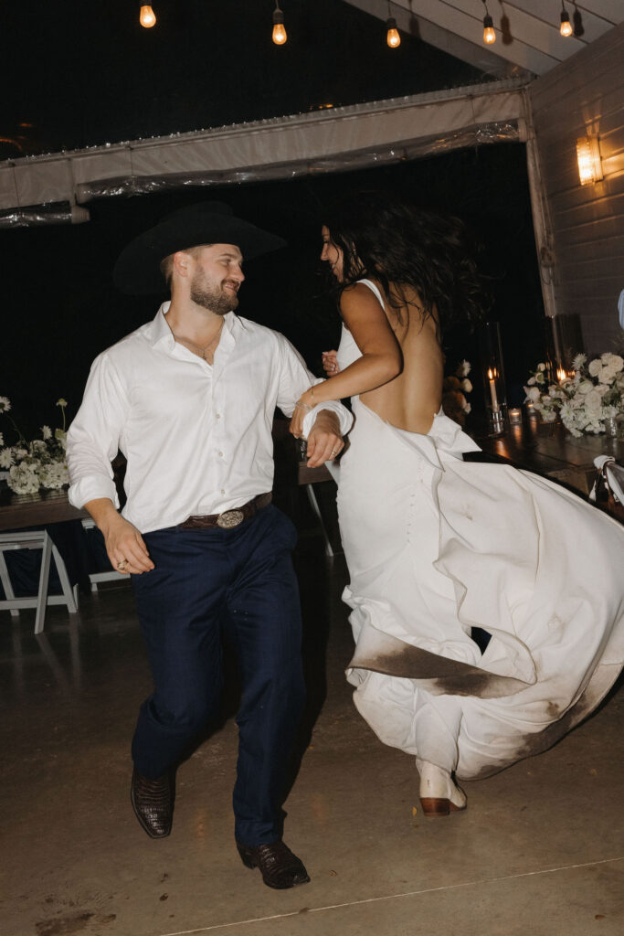 The bride and groom joyfully dance together at the reception, the bride’s dress twirling and showing a playful touch of dirt at the hem.