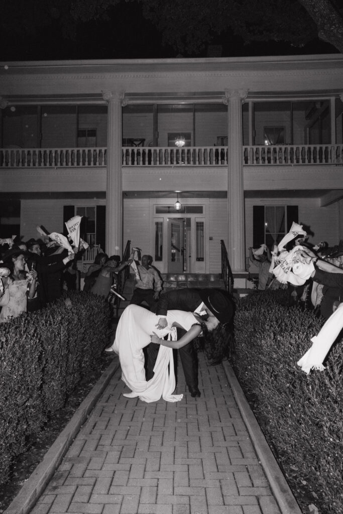 The bride and groom share a dramatic kiss during their send-off, surrounded by guests waving celebratory flags in front of The Winfield Inn.