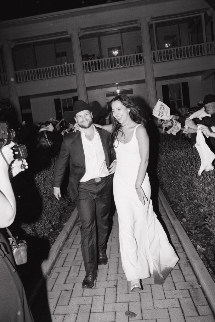 The bride and groom make their grand exit at night, smiling and holding hands as they walk down a brick pathway lined with cheering guests waving custom wedding towels.