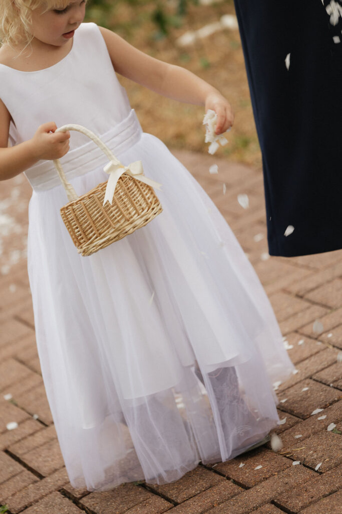 A young flower girl dressed in a white dress is scattering petals along a brick pathway, holding a wicker basket with a cream ribbon tied to it.