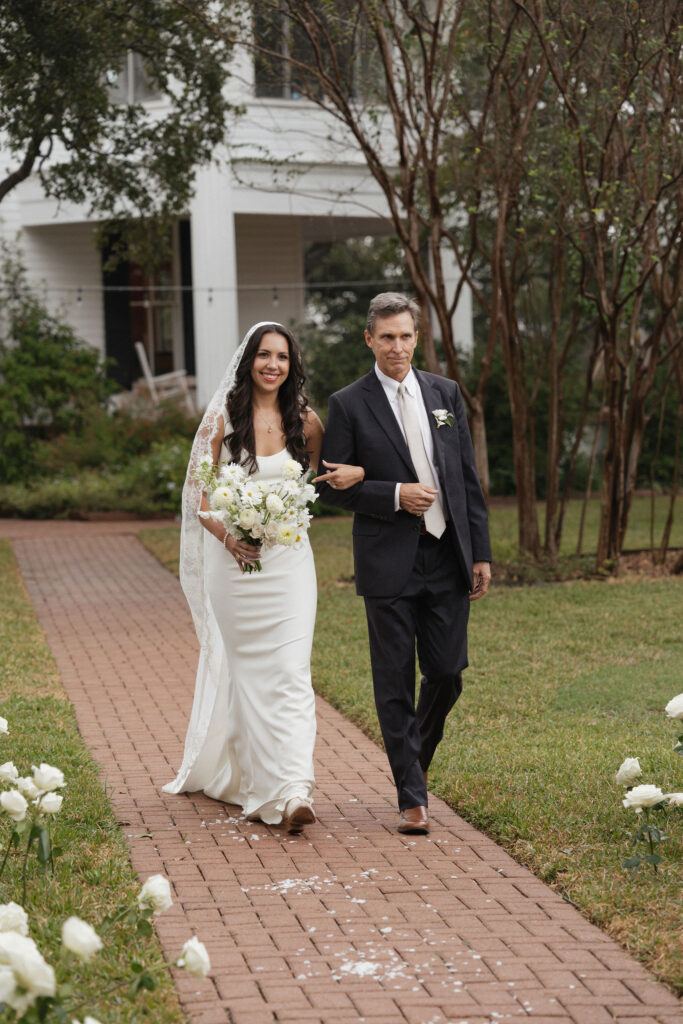 The bride, holding a bouquet of white flowers, walks down the brick aisle arm-in-arm with her father. They are surrounded by guests seated on white chairs amidst a serene outdoor setting.