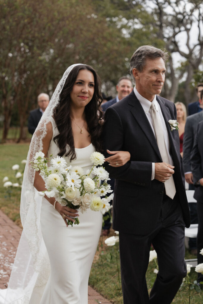 The bride, escorted by her father, walks down the aisle holding a bouquet of white flowers, with a serene smile on her face.