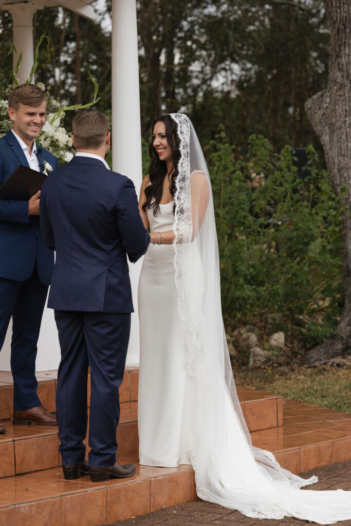 The bride smiles at her groom as they exchange vows in an intimate outdoor ceremony surrounded by lush greenery.