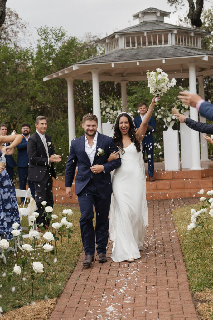 The newlyweds joyfully walk down the aisle after their ceremony, surrounded by cheering guests and white rose arrangements lining the brick path.