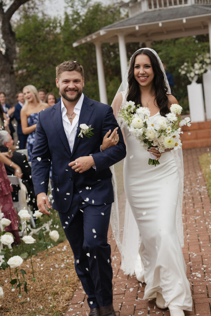 A beaming bride and groom walk down the aisle hand in hand after their ceremony, surrounded by cheering guests and white rose petals.