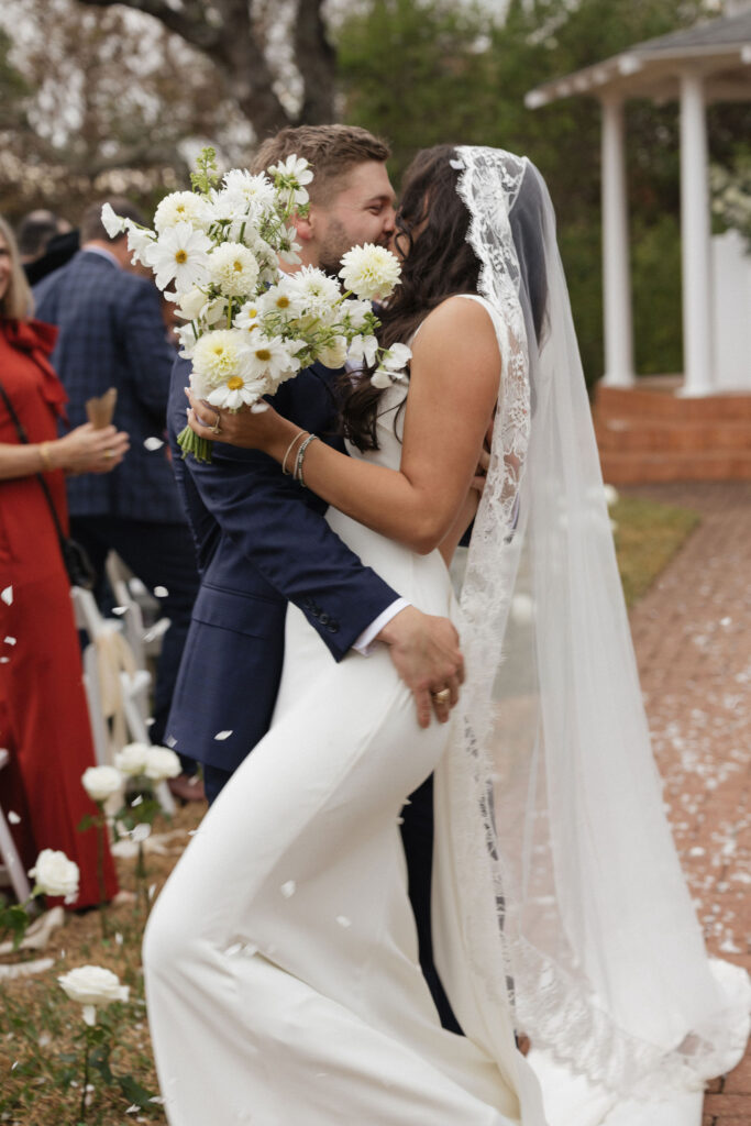 The groom dips the bride for a romantic kiss at the end of the aisle, with guests cheering in the background.