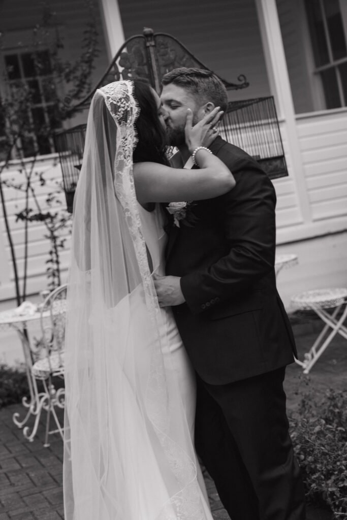 The bride and groom share an intimate kiss under the bride’s lace veil, with the historic white house of The Winfield Inn as a charming backdrop.