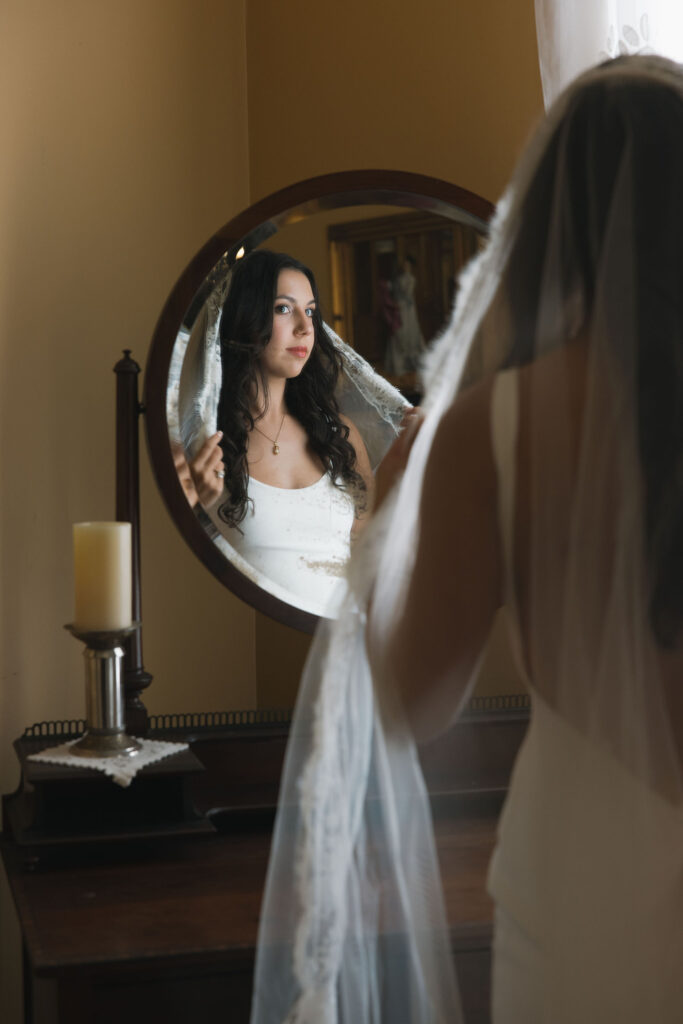 A bride looks into a vintage mirror, adjusting her veil, her expression calm and serene in the warm light of The Winfield Inn.