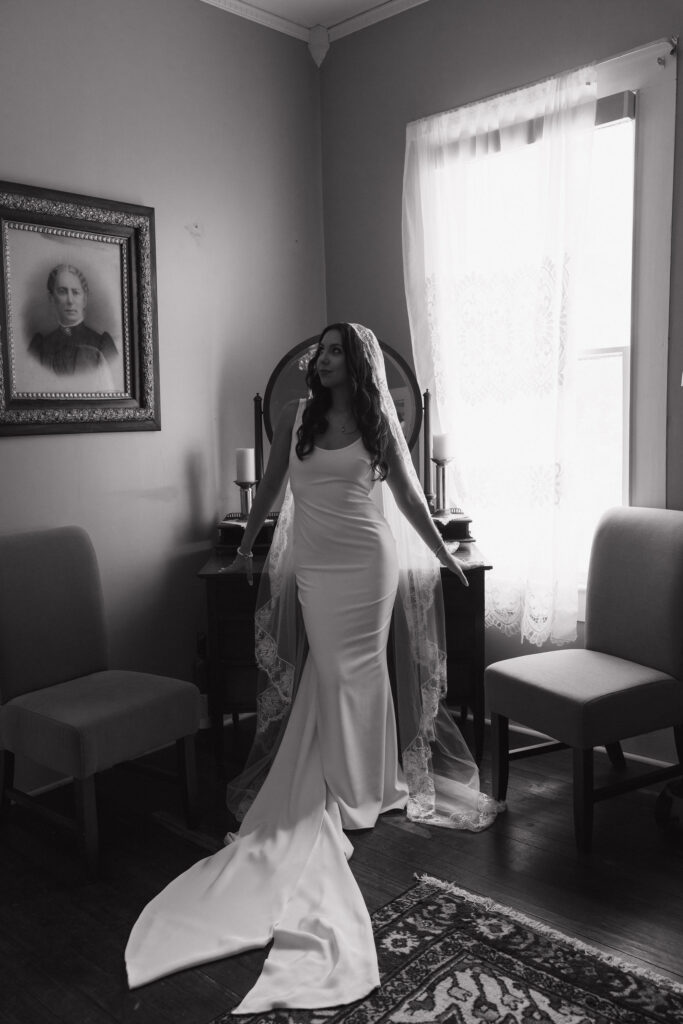 The bride stands in a softly lit room, wearing her wedding gown and veil, next to antique furniture and a vintage portrait on the wall.