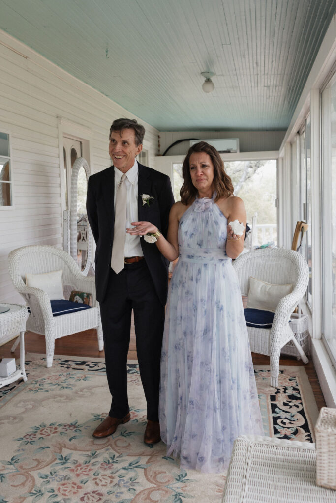 The bride’s parents stand on a charming porch with wicker furniture, the father in a black suit and the mother in a flowing floral gown, holding back tears of joy.