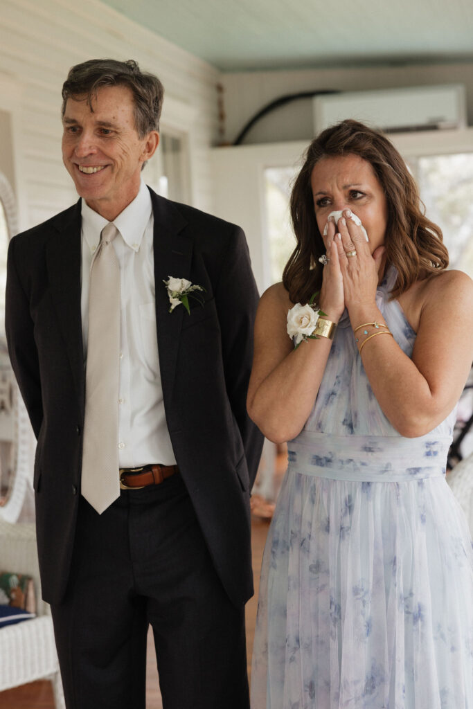 The bride’s parents watch with emotional expressions during a touching moment of the wedding day, standing side by side.