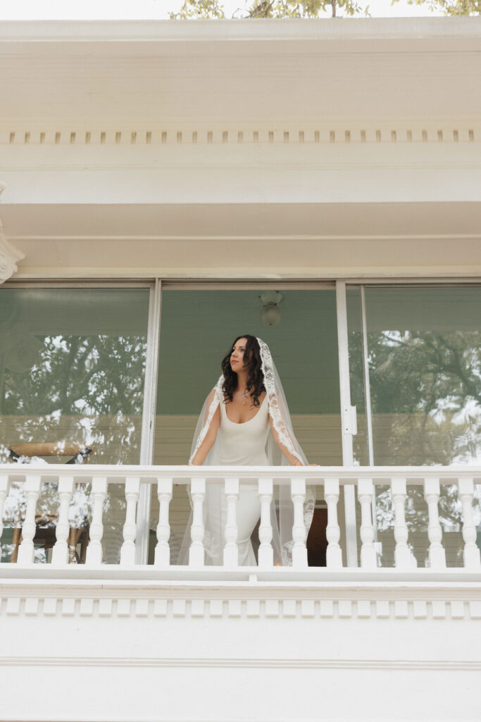 The bride poses on a white balcony, her lace veil flowing as she gazes into the distance, with soft natural light enhancing the scene.