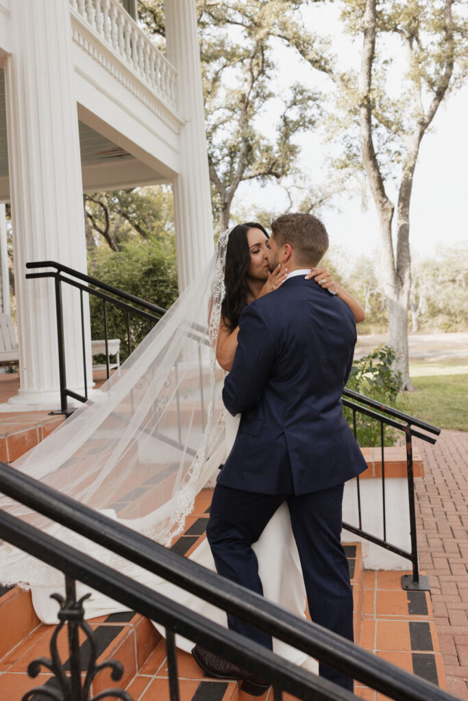 The bride and groom share a tender kiss on the steps of the inn, with the bride’s lace veil cascading down the brick stairs, surrounded by lush greenery.