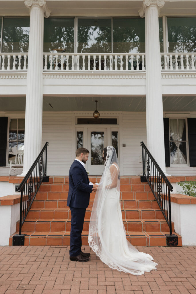 A couple exchanging vows on the steps of a charming white colonial-style mansion, with the bride wearing a long veil and the groom in a navy suit.