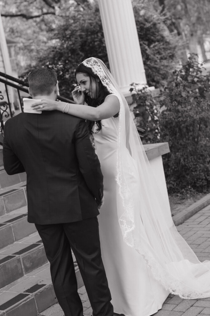  The bride approaches the groom outdoors, her lace veil flowing as she carries her bouquet, and he waits in a cowboy hat, grinning.