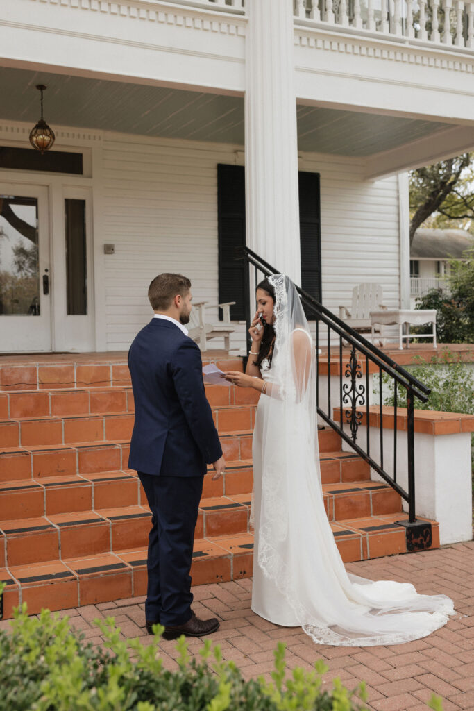 The bride, standing at the base of the inn’s staircase, holds her vows in one hand, visibly emotional as she reads to the groom.