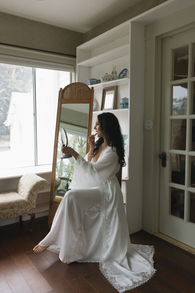 The bride applies lipstick while sitting gracefully in a vintage chair, surrounded by the cozy interiors of The Winfield Inn.