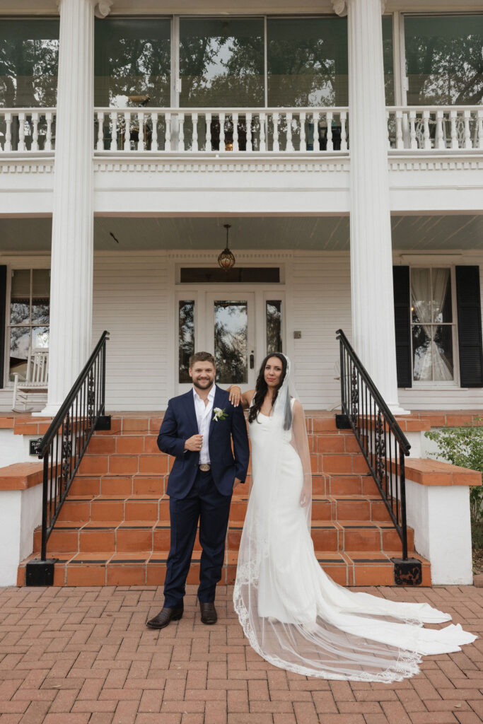 The bride, wearing a flowing white gown with a lace veil, meets her groom on the steps of The Winfield Inn’s grand porch for a special moment.