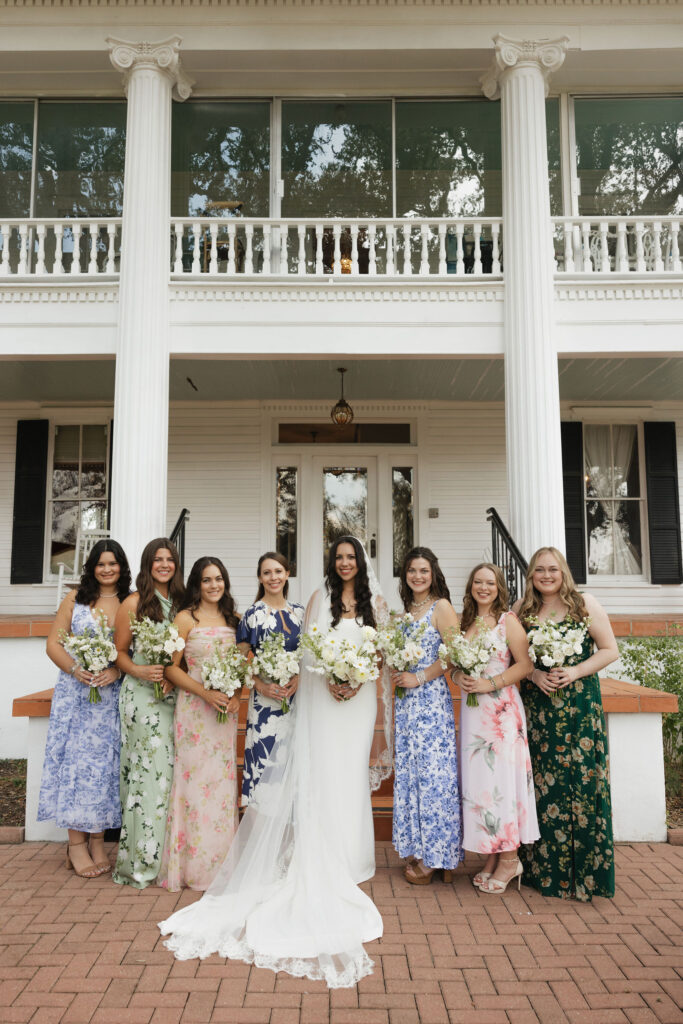 The bride, in a classic white gown and veil, stands alongside her bridesmaids dressed in floral-patterned dresses, holding white bouquets in front of the inn's elegant columns.