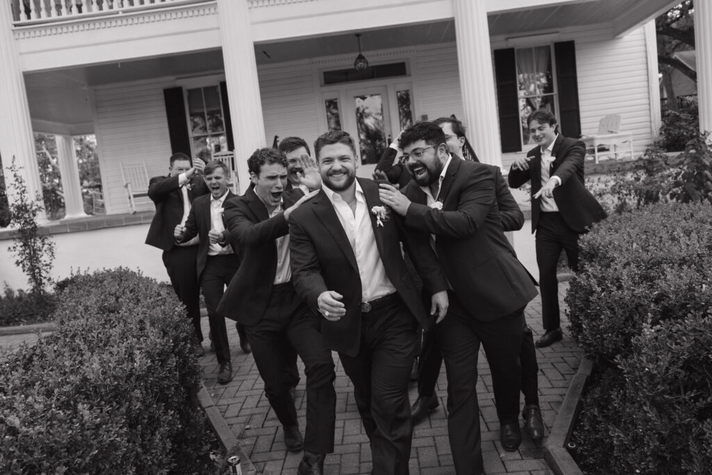 A candid black-and-white shot of the groom and his groomsmen laughing and cheering in front of The Winfield Inn’s porch.