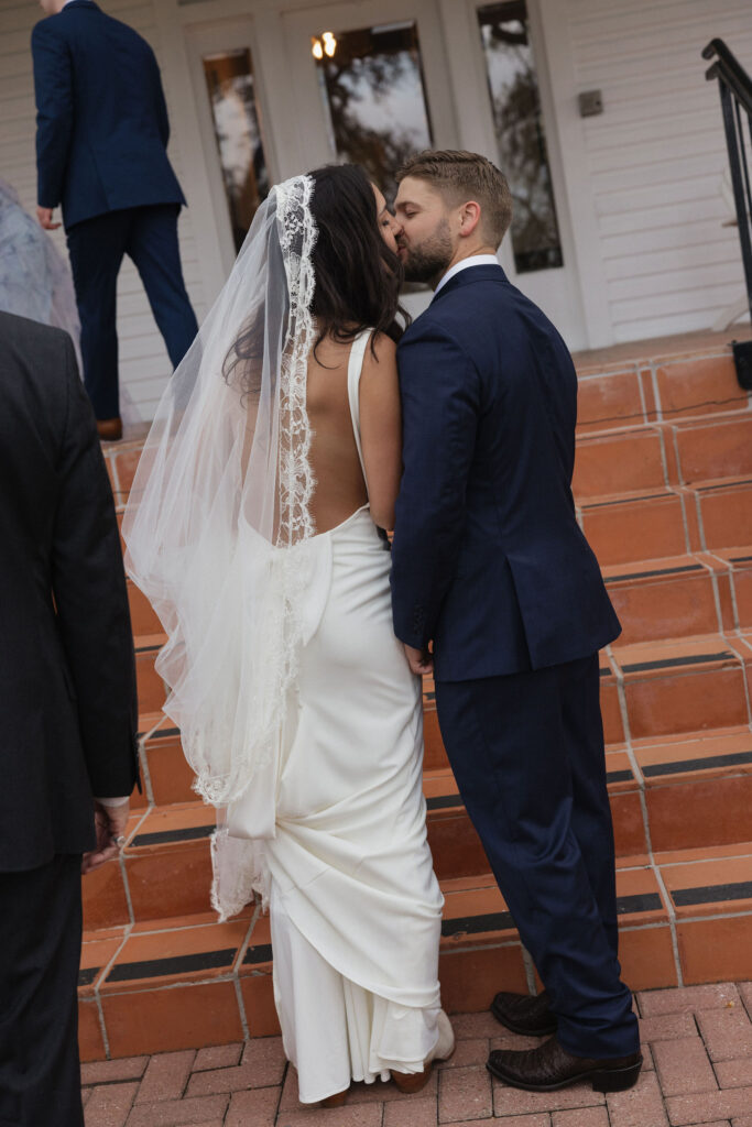 The bride and groom share a quiet kiss on the steps of the mansion after their ceremony, showcasing her elegant open-back gown and lace veil.