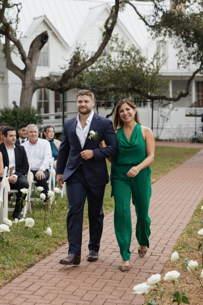 The groom walks down the aisle with his mother, who wears an elegant green jumpsuit, smiling warmly as guests look on.