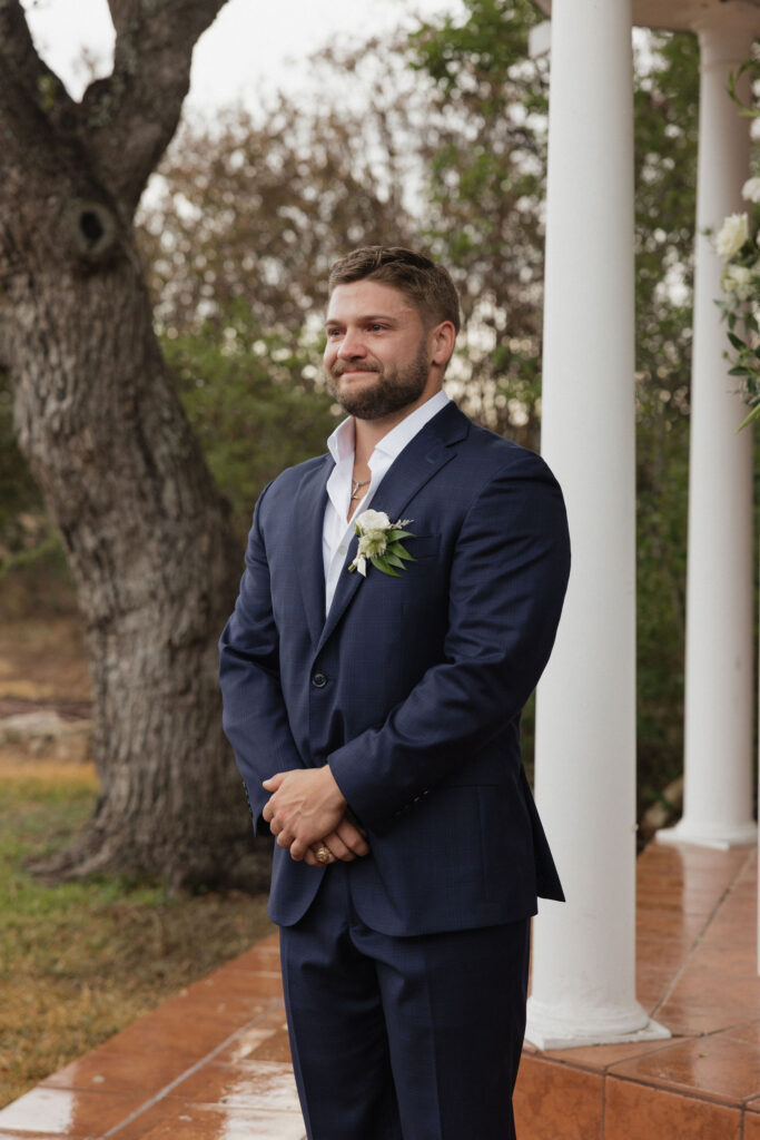 The groom stands patiently in a navy suit with a white boutonniere, smiling softly as he waits for his bride.