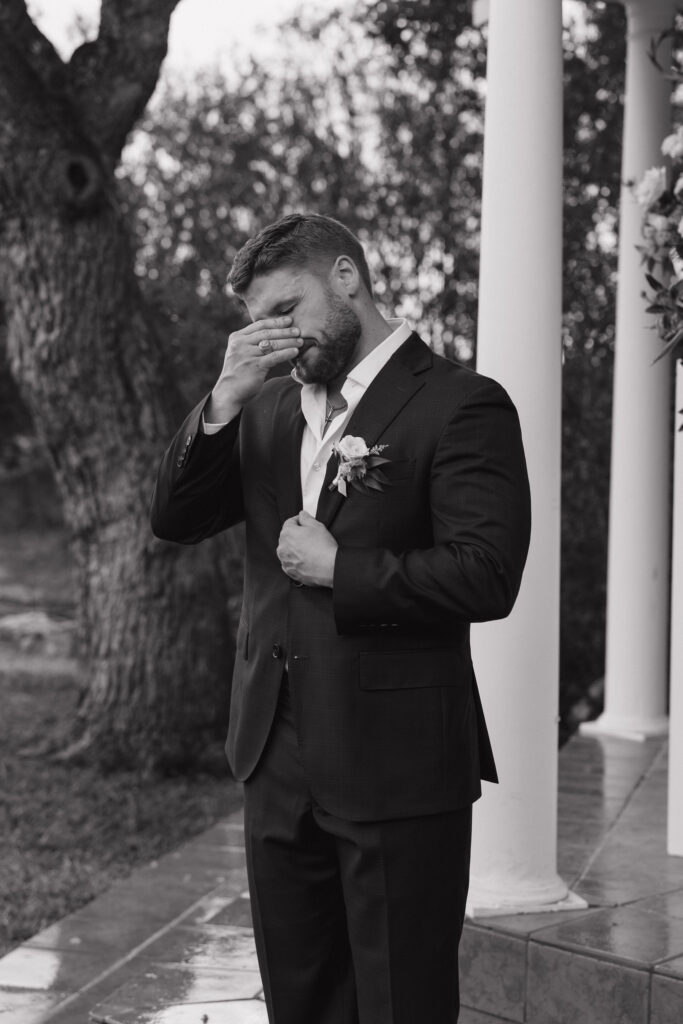 The groom wipes away tears during the ceremony, standing under the gazebo surrounded by oak trees.