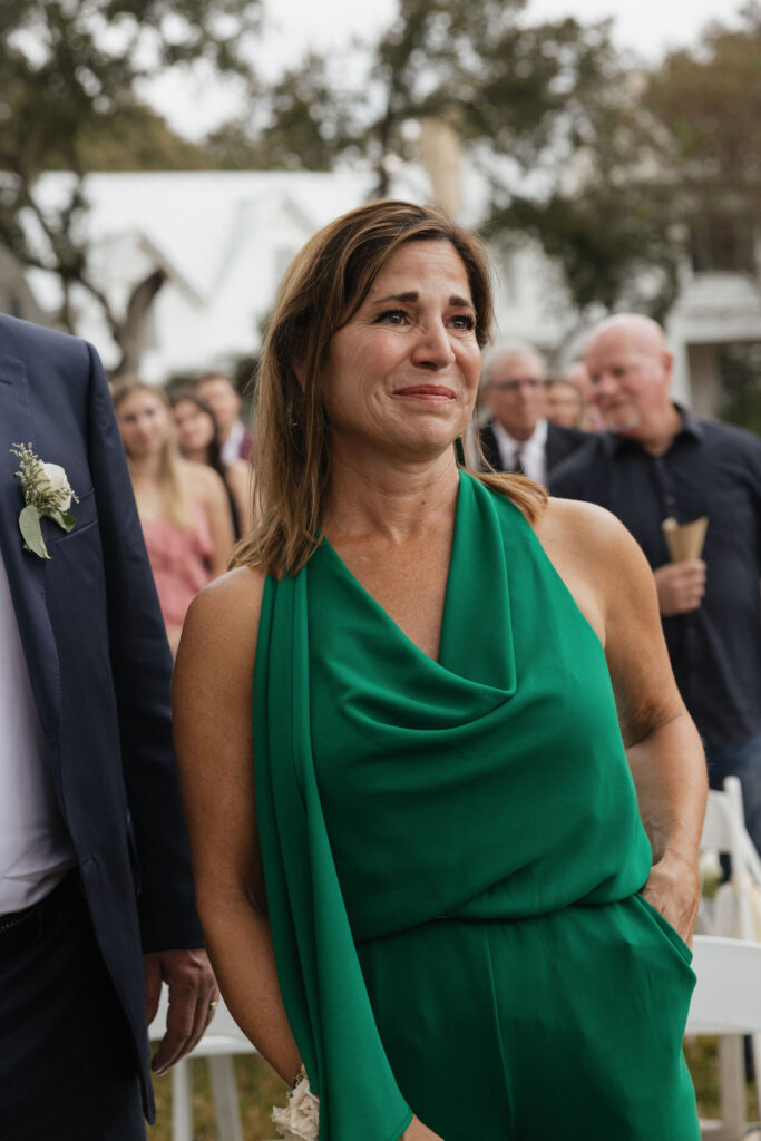 A close-up of the groom's mother in a green dress, wiping away tears of joy during the ceremony, with guests in the background.