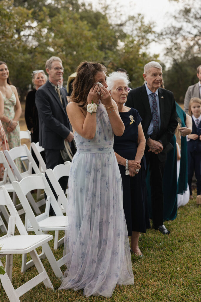 A mother of the bride wipes tears of joy during the ceremony while standing amidst seated and standing wedding guests.