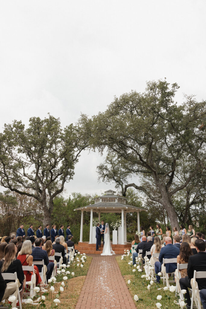 A wide view of the ceremony under the venue’s gazebo, with white flowers lining the brick aisle and guests seated on either side.