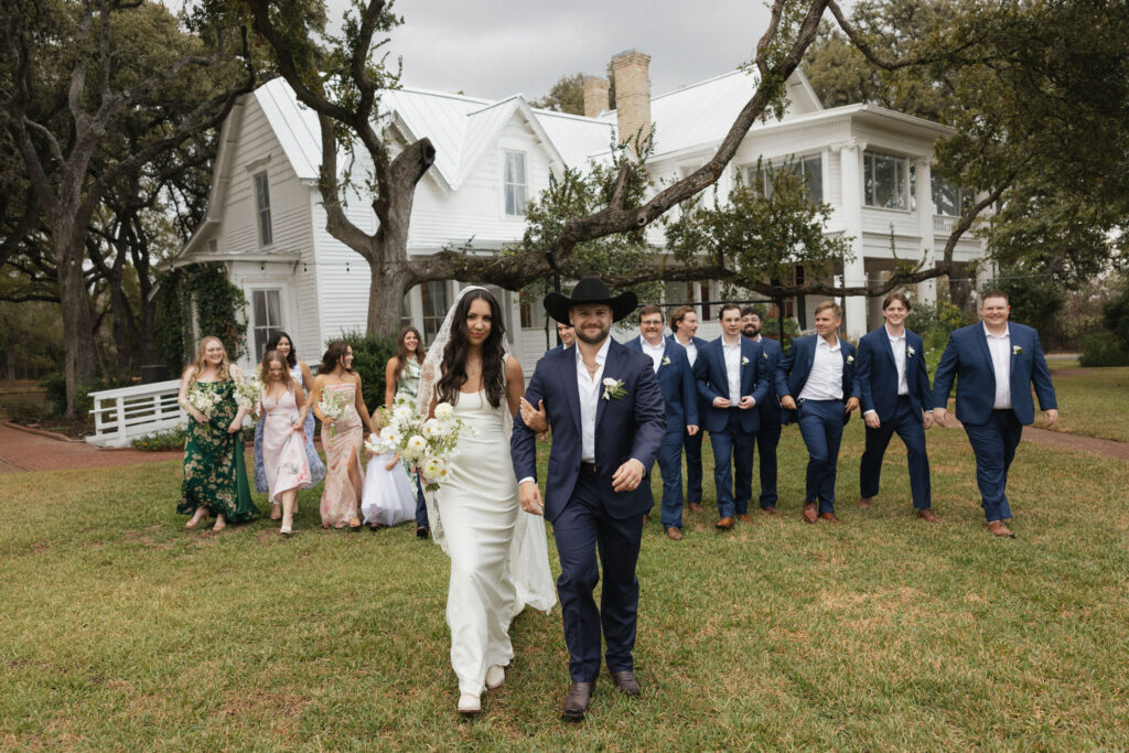 The bride and groom lead their wedding party across a lawn, the historic white mansion behind them, as bridesmaids in pastel dresses and groomsmen in navy suits follow.