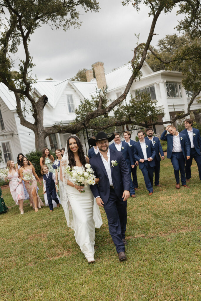 The bride and groom lead their wedding party across a lawn, the historic white mansion behind them, as bridesmaids in pastel dresses and groomsmen in navy suits follow.