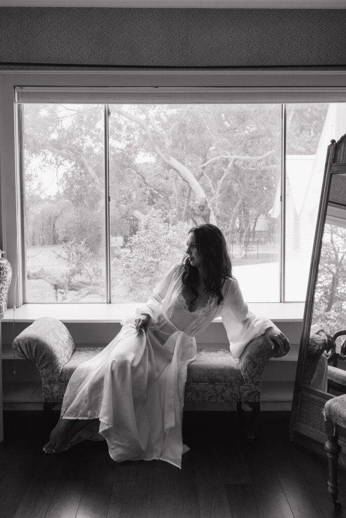 The bride sits gracefully on a chaise lounge by a large window, looking outside at the scenic grounds of The Winfield Inn.