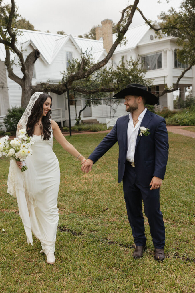 The bride and groom share a lighthearted moment in the garden, with the groom wearing a cowboy hat and the bride holding a bouquet of white flowers.