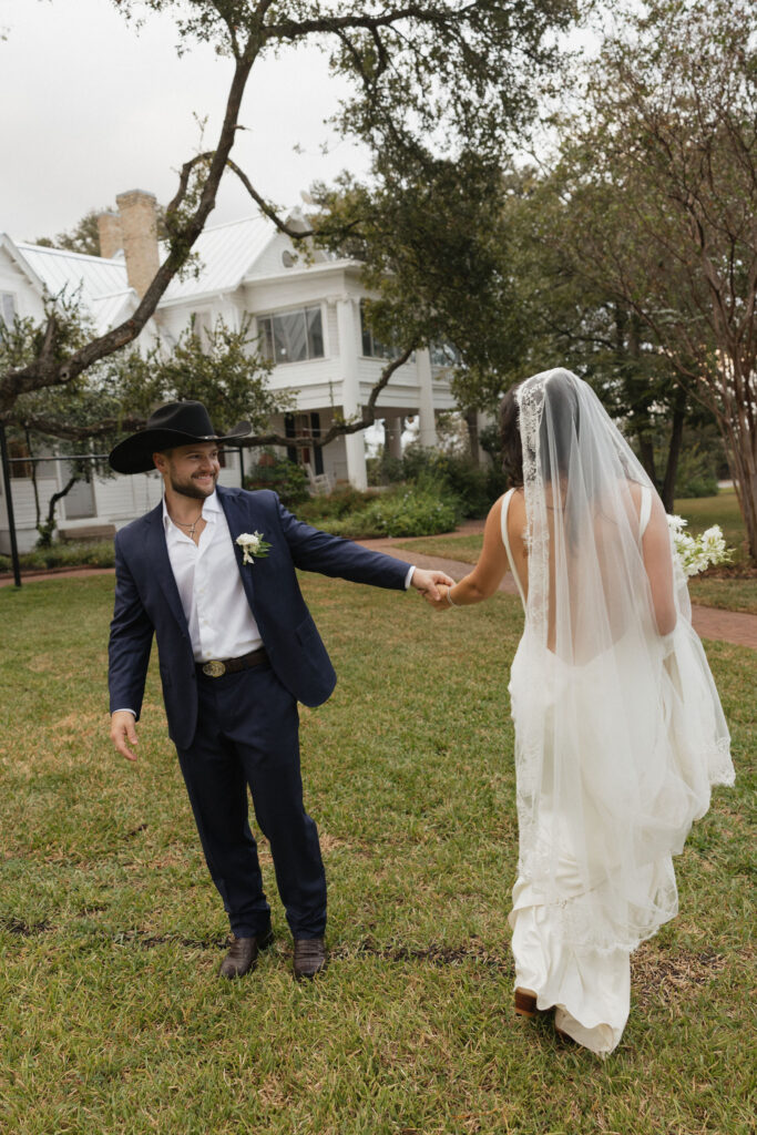 The groom, dressed in a navy suit and cowboy hat, smiles warmly as he gently holds the bride's hand, leading her across the lawn in front of the historic Winfield Inn.